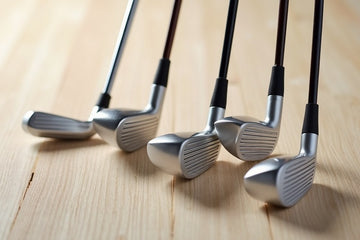 Five silver and black golf iron clubs arranged diagonally on a light wood surface.