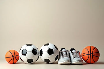 A collection of sports equipment arranged in a row against a solid beige background, including two orange basketballs, two black and white soccer balls, and a pair of white athletic sneakers.