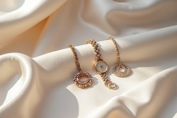 Three gold-toned jewelry pieces arranged on draped cream satin fabric: a bracelet, a watch with a round dial, and an open circular pendant. The jewelry has a warm gold color with clear stones.